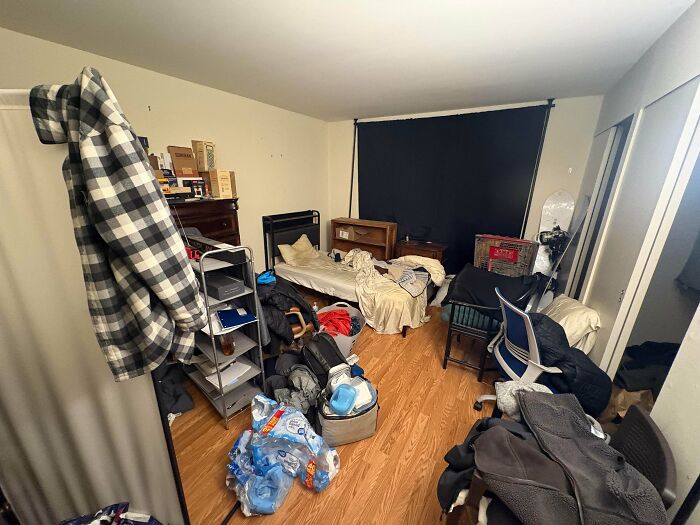A cluttered male living space with clothes, bottles of water, and various items strewn across the floor and furniture.