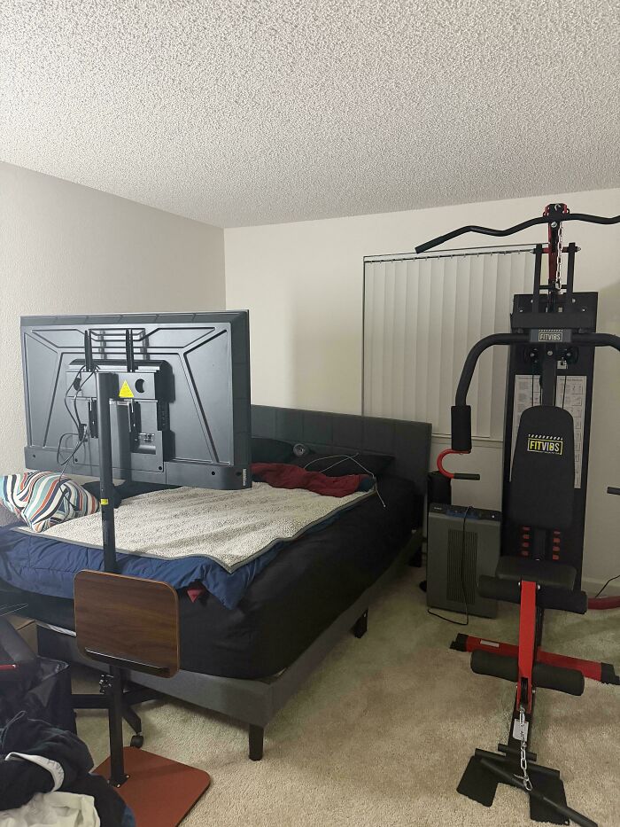 A neatly organized male living space with a large TV mounted above the bed and a multi-gym machine.