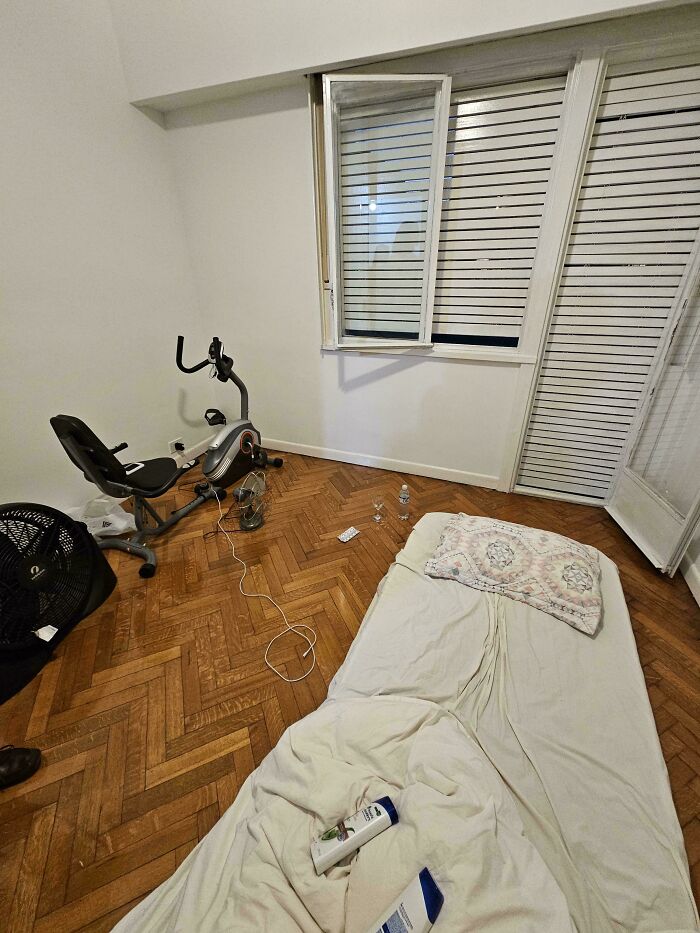 A minimalist male living space featuring a floor mattress, an exercise bike, and a fan on a hardwood floor.