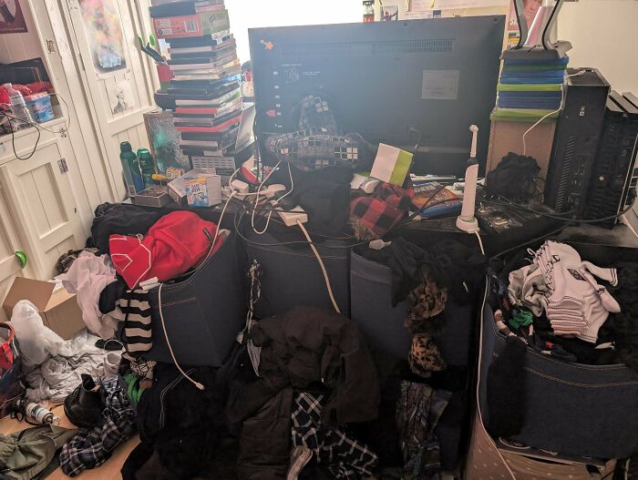 An extremely disheveled male living space with piles of clothes, books, and electronics covering almost every surface.