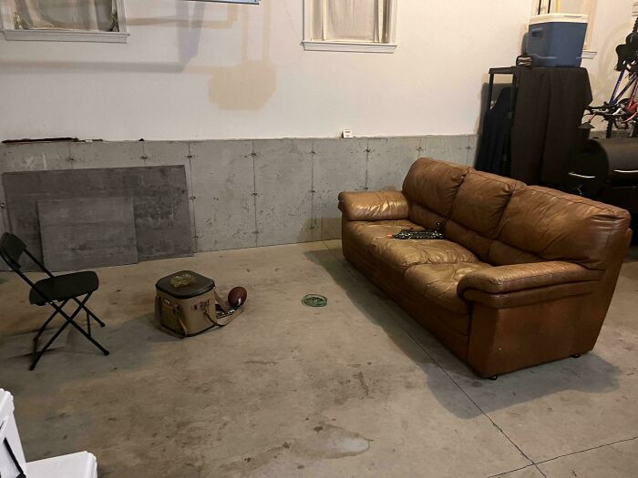 A garage transformed into a male living space with a worn leather couch, a folding chair, and a cooler on a concrete floor.