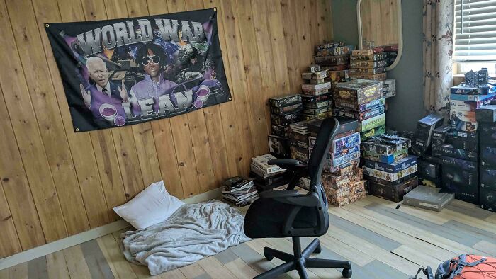 A male living space featuring a large collection of board games stacked against a wood-paneled wall, with a small bed on the floor.