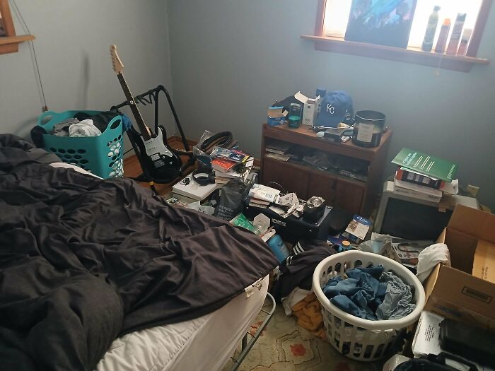 A very untidy male living space with clothes, boxes, and a guitar, suggesting disorganization.
