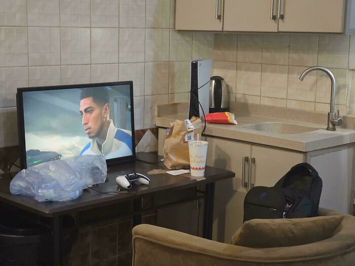 A cluttered male living space with a TV, game console, and fast food near a kitchen sink.