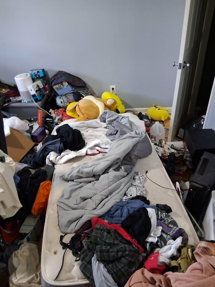 A very cluttered male living space with clothes, items, and a pillow scattered across a mattress on the floor.