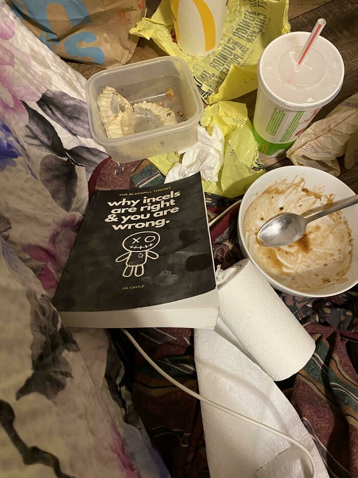 A close-up of a chaotic male living space with fast-food wrappers, a dirty bowl, and a book on a crumpled blanket.