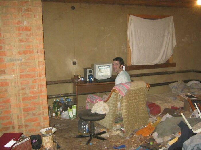 A male smiling at a computer in a sparsely furnished, untidy room with a brick wall, reflecting on male living spaces.