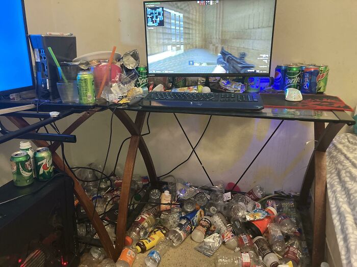 A messy male living space with a computer, soda cans, and trash covering the desk and floor.