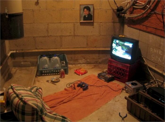 A cluttered male living space in a basement with a small TV, a picture on the wall, and basic furniture.