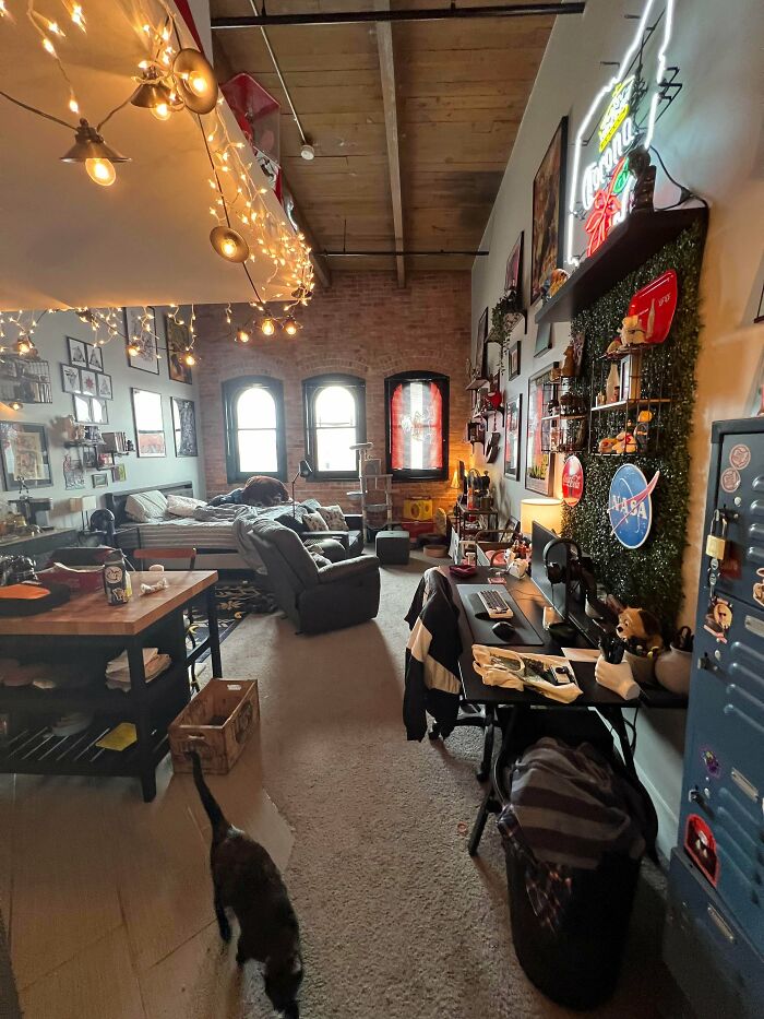 A cozy and eclectic male living space with string lights, brick walls, multiple framed pictures, and a home office setup.