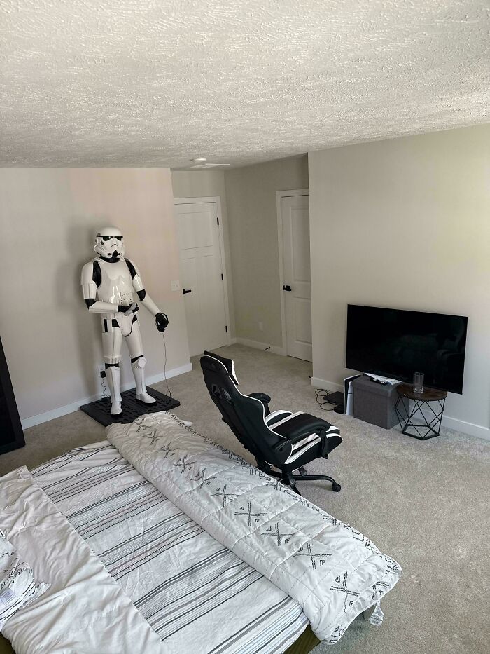 A bedroom in a male living space with a Stormtrooper figure, a gaming chair, a bed with striped bedding, and a wall-mounted TV.