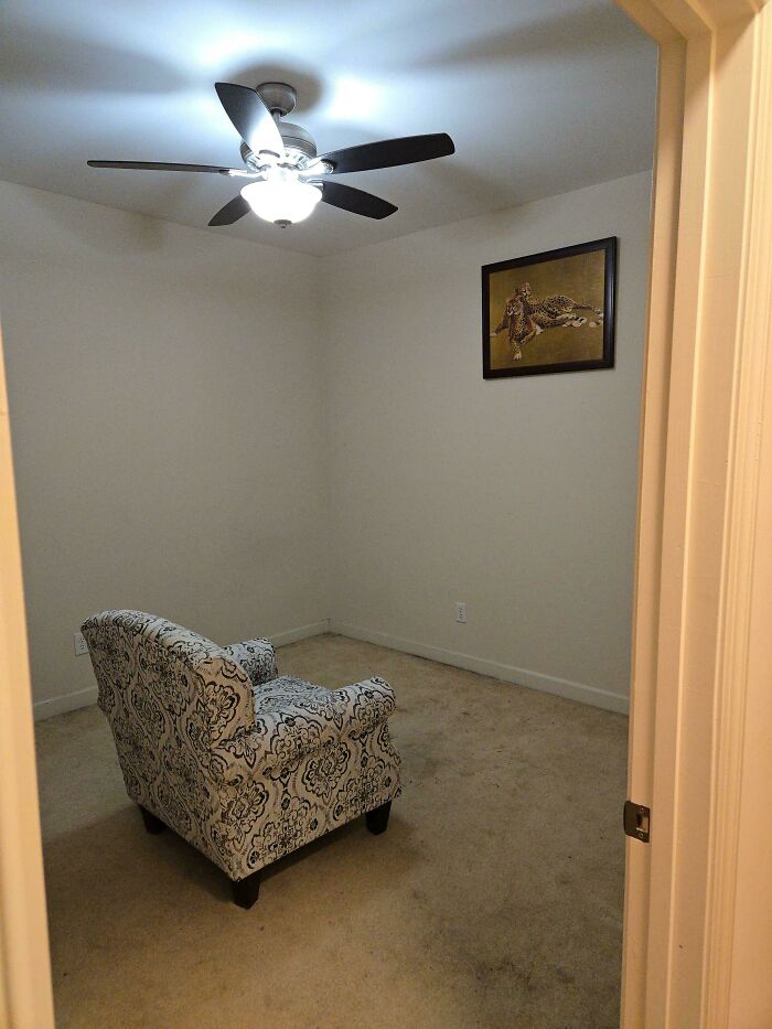 A minimalist male living space with a single patterned armchair, a ceiling fan, and a framed picture on a plain wall.