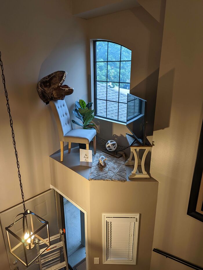 An elevated male living space nook with a dinosaur head, a chair, and a plant near a large window, overlooking a lower area.