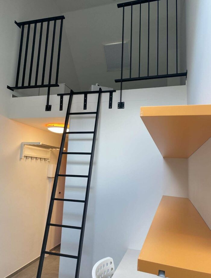 A modern male living space with a black ladder leading to a loft, black railings, and an orange shelf.