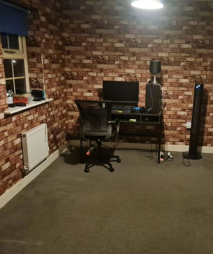 A simple male living space featuring a brick-patterned wall, a desk with a computer setup, and a floor fan.