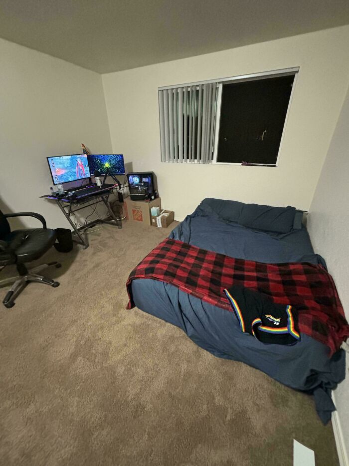 A cozy male living space featuring a bed with a red plaid blanket and a gaming setup with dual monitors near a window.
