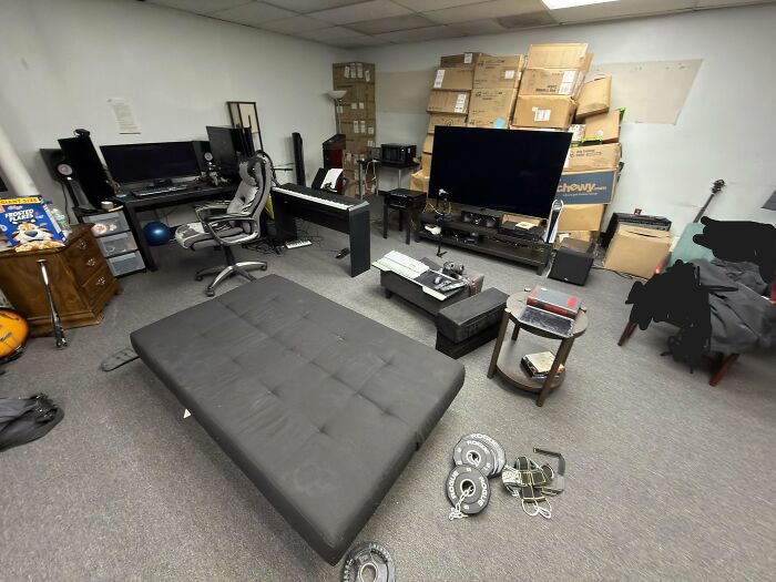 A male living space used for various activities, including a futon, computer desk, piano, and stacks of boxes.