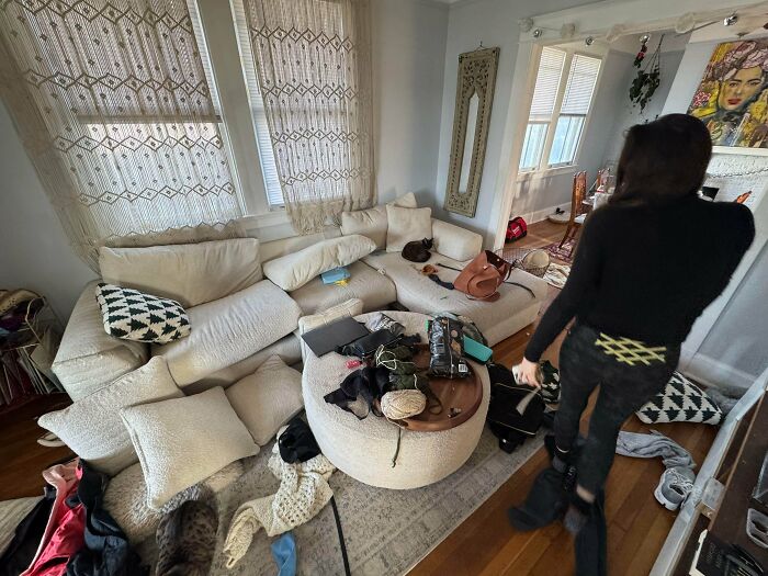 A messy male living space with a white sectional sofa, a round coffee table, and various items scattered around the room.