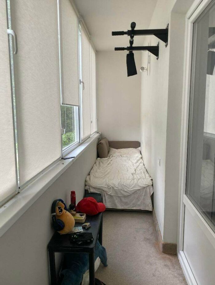 A small, narrow male living space on a balcony with a bed, small table, and a pull-up bar.
