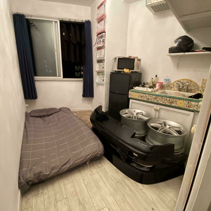 A small, cluttered male living space with a bed, a refrigerator, a sink, and car rims stored indoors.