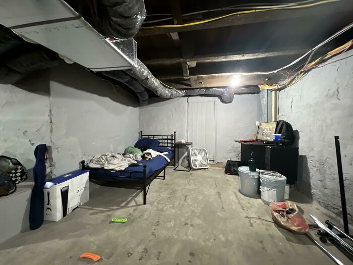 A rough male living space in a basement with exposed pipes, a bed, a desk, and several buckets.