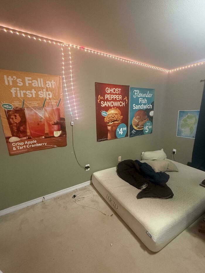 A messy male living space with a mattress on the floor, string lights, and fast-food posters on the wall.