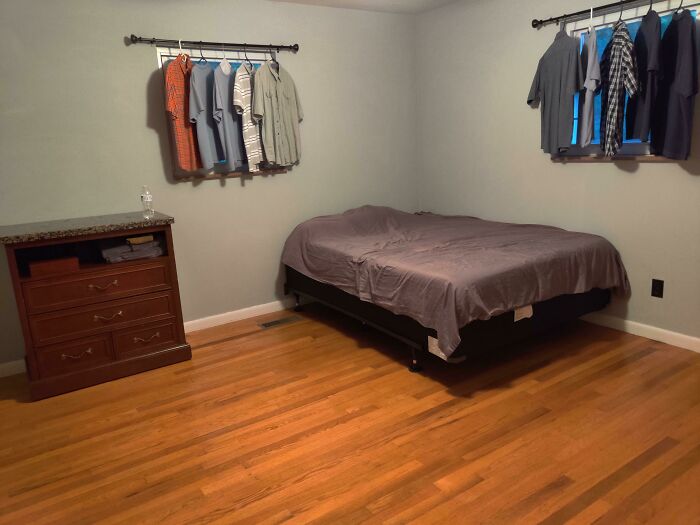 A sparsely furnished male living space with a bed and shirts hanging in windows, showing how not to live.