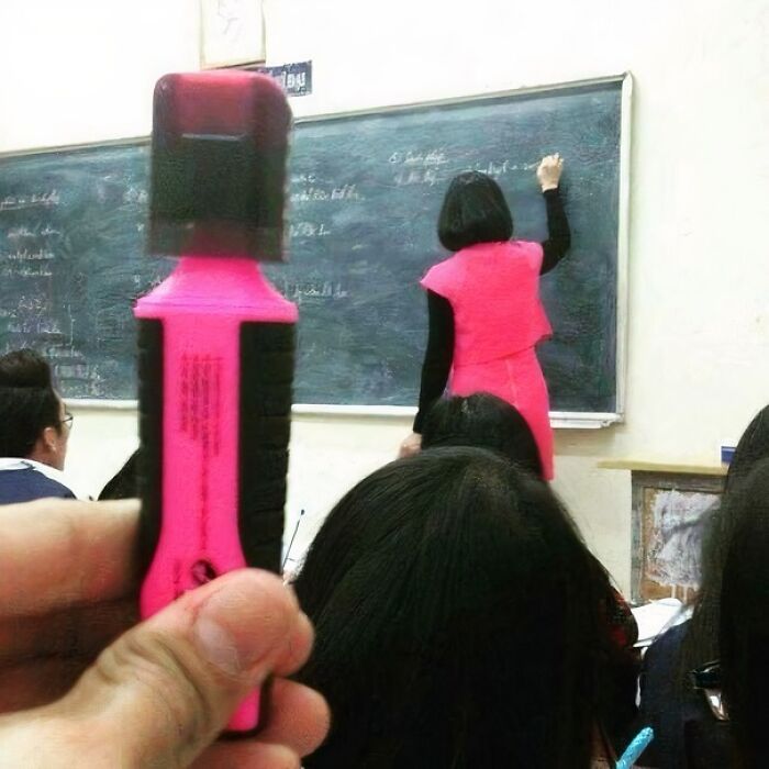 A person holding a pink marker in the foreground, perfectly aligned with a teacher in a matching pink dress writing on a blackboard. Cannot Explain Pics.