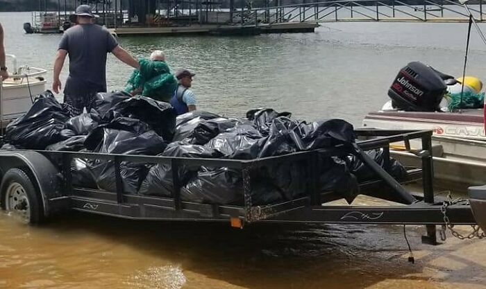 A trailer full of black trash bags in the water, with people and boats around, as trash takes over the world.