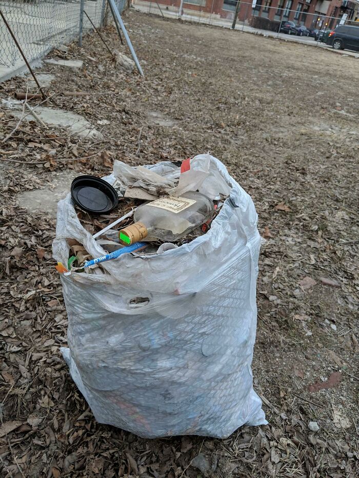 A white plastic bag filled with various trash items, including bottles and plastic, sits on a ground covered with d**d leaves, showing the world's trash.