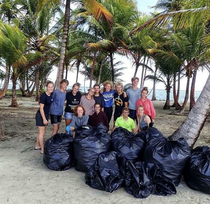 A group of people standing on a beach with palm trees and bags of collected trash, taking over the world.