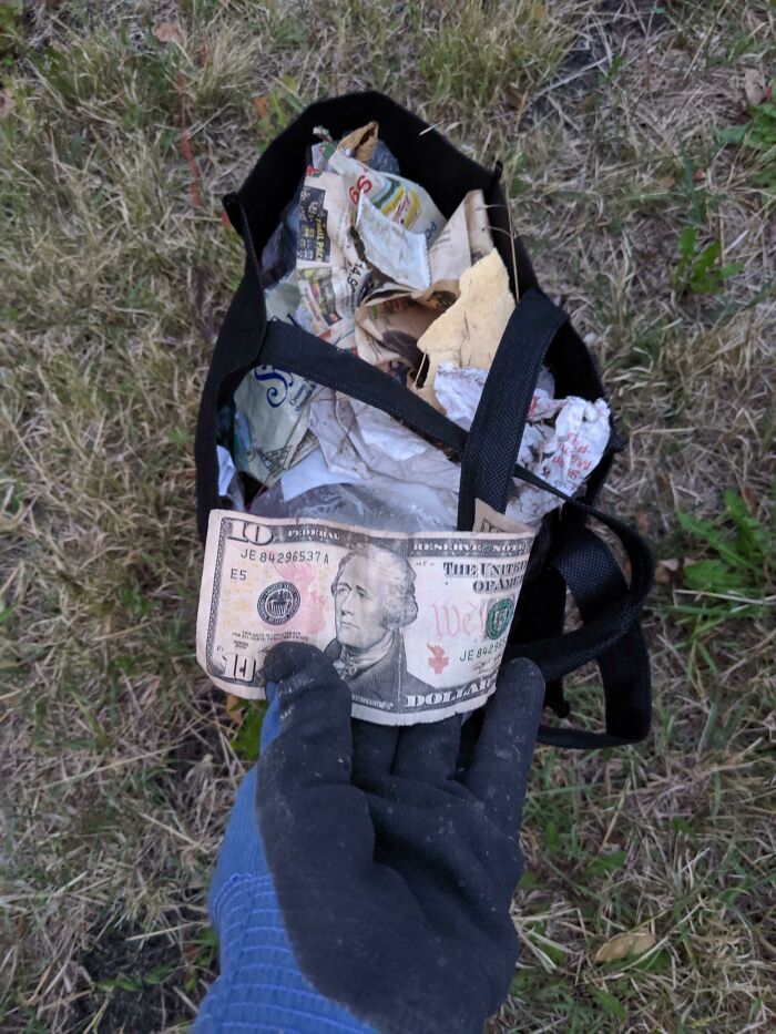 A gloved hand holding a ten-dollar bill, with a bag full of trash in the background, highlighting the issue of trash.