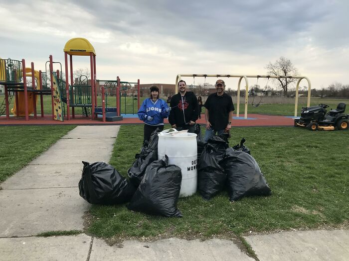 Three people standing with black trash bags and a white barrel in a park, after collecting trash.