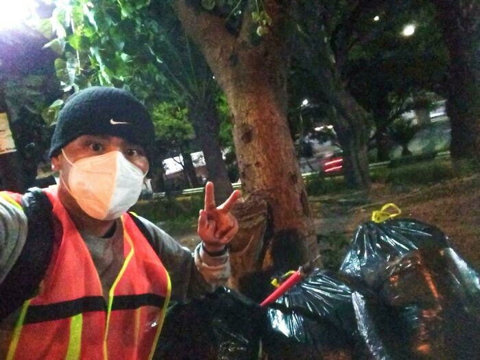 A person in a mask and reflective vest poses with bags of trash, acting as an influencer for cleaning up trash.