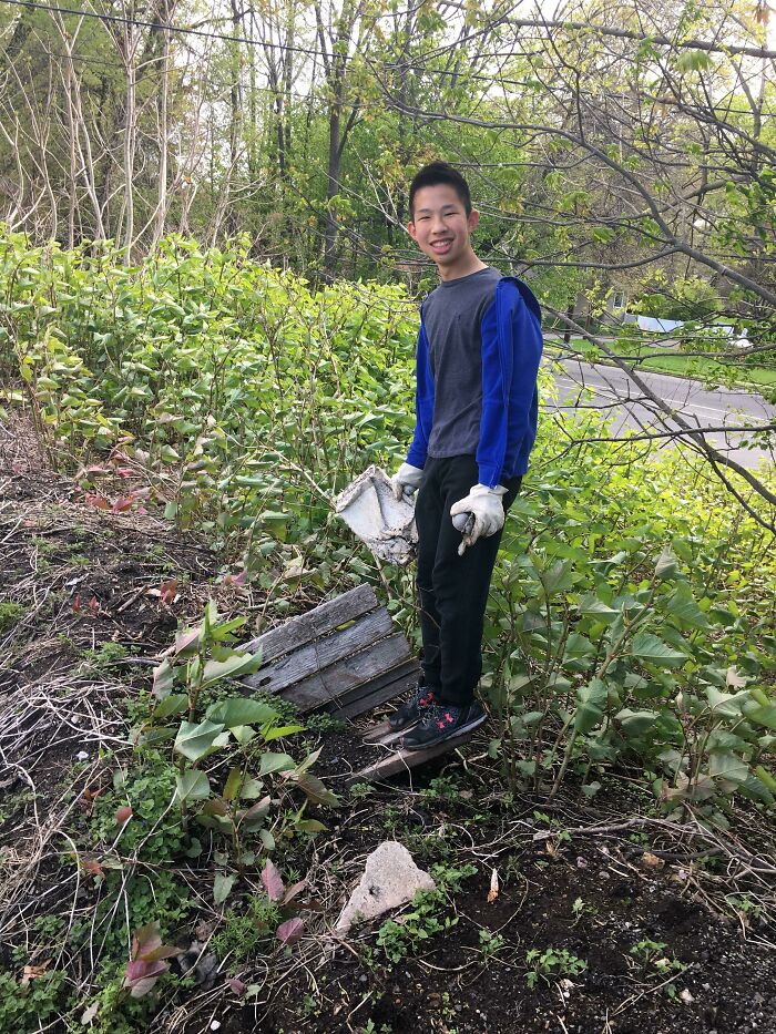 A young person smiles while standing amidst overgrown foliage, holding a piece of trash, having picked up trash.