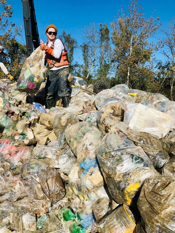 A person stands on a massive pile of clear trash bags filled with waste, under a bright blue sky.