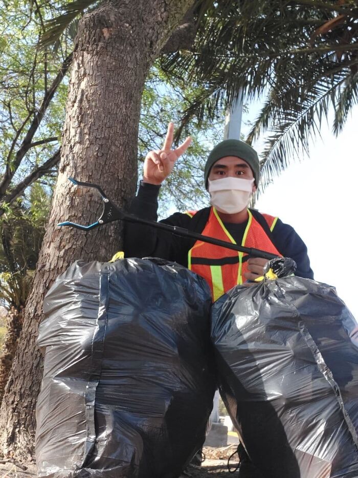 A person in a mask and safety vest poses with a trash picker and two full bags of trash, showing peace sign.