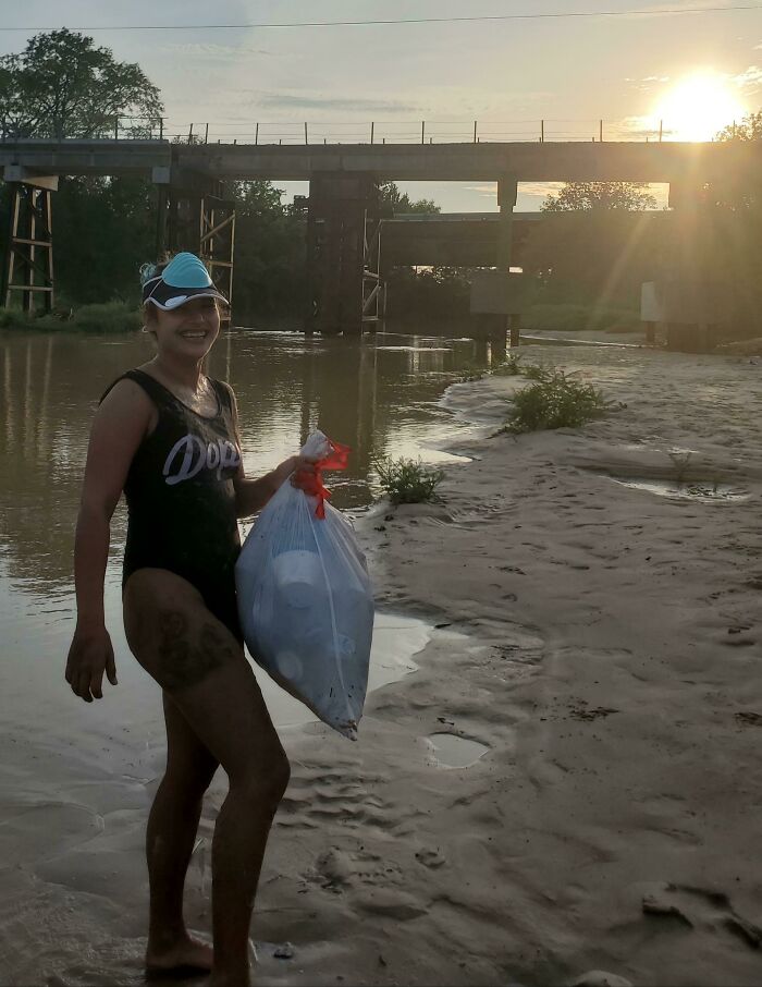 A smiling woman stands in a shallow river with a bag of trash she collected, sunset in the background.