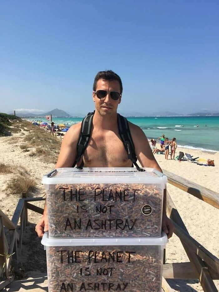 A shirtless man on a beach holding two clear bins filled with cigarette butts. People are taking over the world.