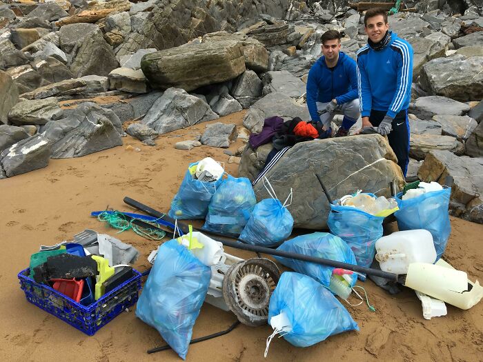 Two young men on a sandy beach next to many bags of collected trash and debris, after cleaning up the world.