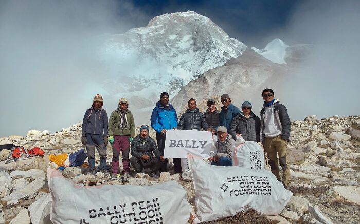 A group of people with bags labeled Bally Peak Outlook Foundation stand in front of a snowy mountain, showing people cleaning up trash.