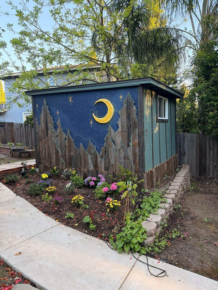 An exterior DIY home project transforming a shed with a creative mural featuring a crescent moon and mountains, surrounded by flowers.