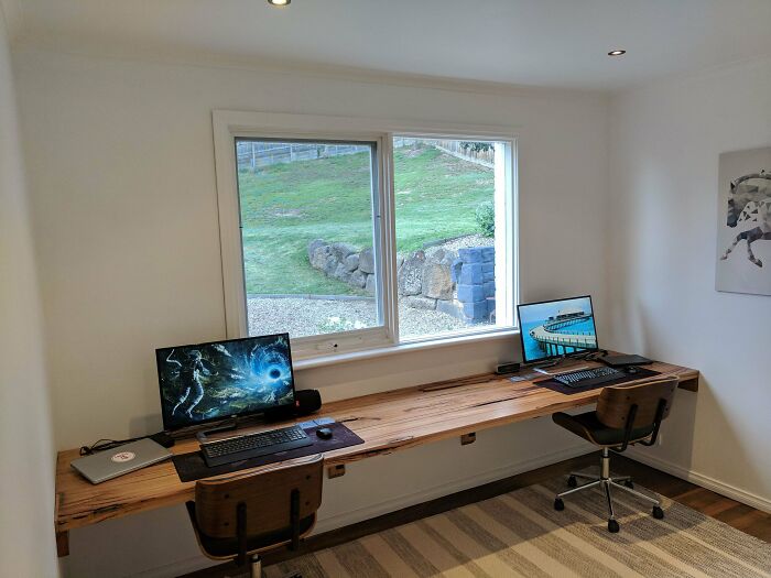 A DIY home project featuring a long wooden desk with two computer setups in a bright room overlooking a green landscape.