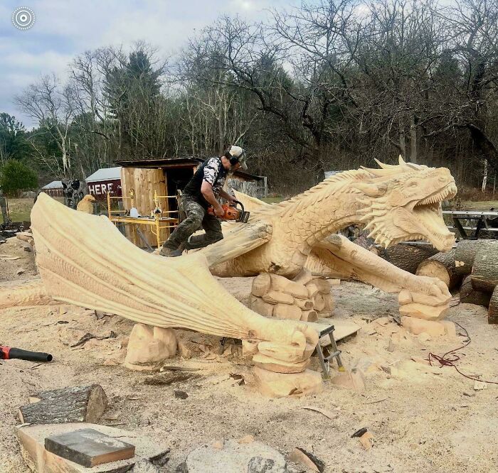 A craftsman uses a chainsaw to carve a detailed wooden dragon sculpture, showcasing a wonderful and weird creation.