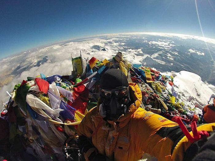 Wild pictures: A person in a yellow jacket and oxygen mask on a snowy mountain peak adorned with prayer flags, appreciating the wonderful world.