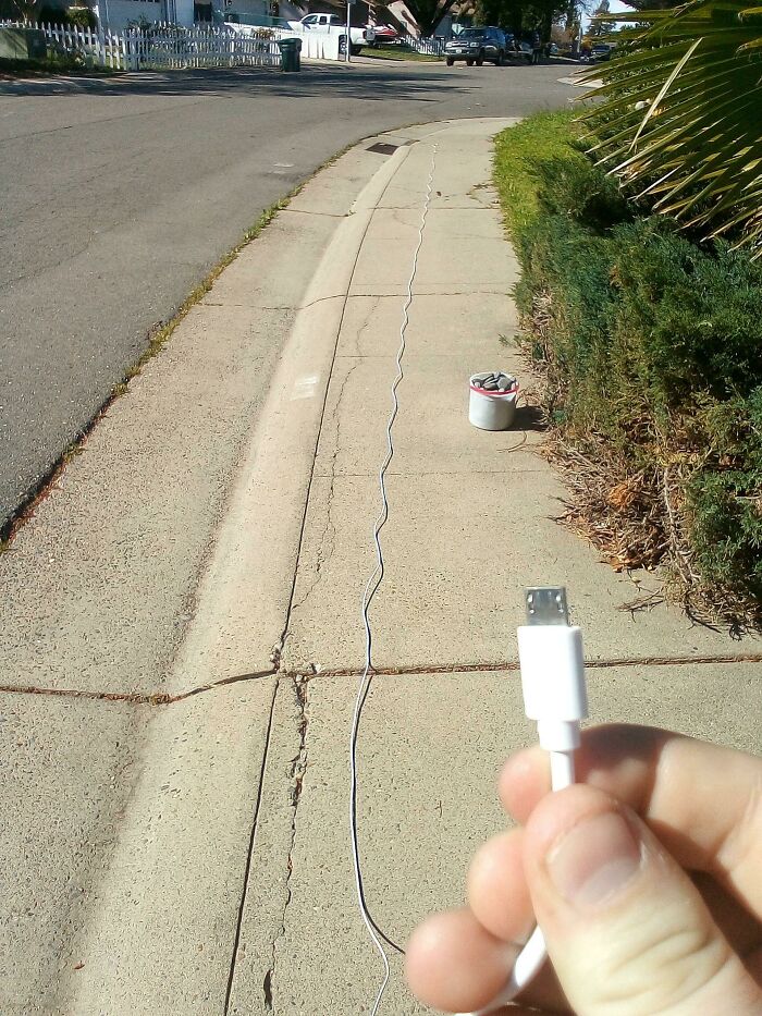 A hand holding a USB cable, extending down a sidewalk towards a bucket, a wonderful and weird outdoor charging setup.