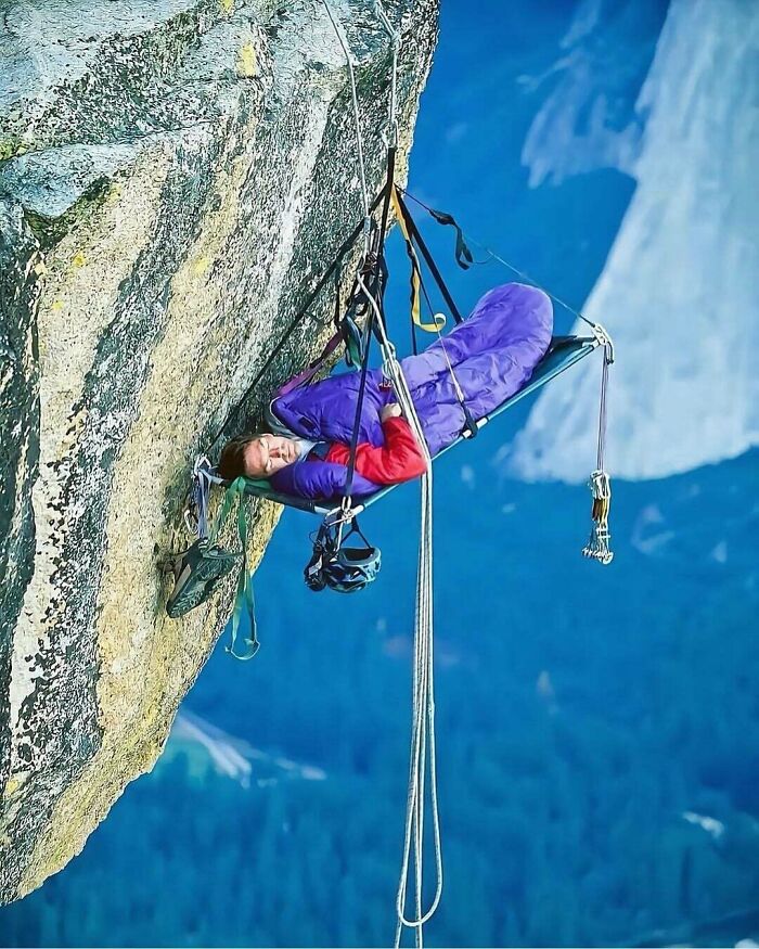 A climber in a purple sleeping bag suspended on a portaledge on a sheer rock face, a wonderfully weird sleeping arrangement.