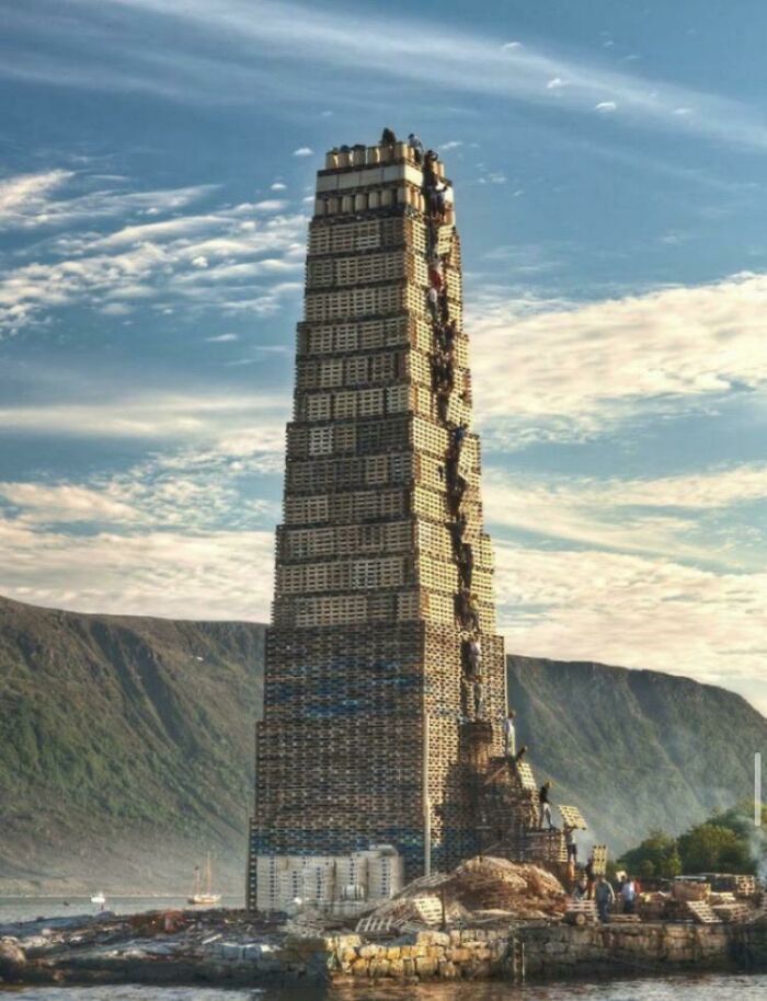 A towering structure built from wooden pallets on a rocky outcrop near a body of water, with mountains in the background, a wild picture of the world.