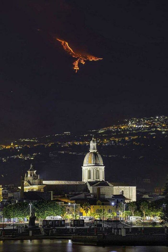A night-time view of a city with a domed building in the foreground and a fiery, dragon-like lava flow on a distant mountain, a wonderful world.