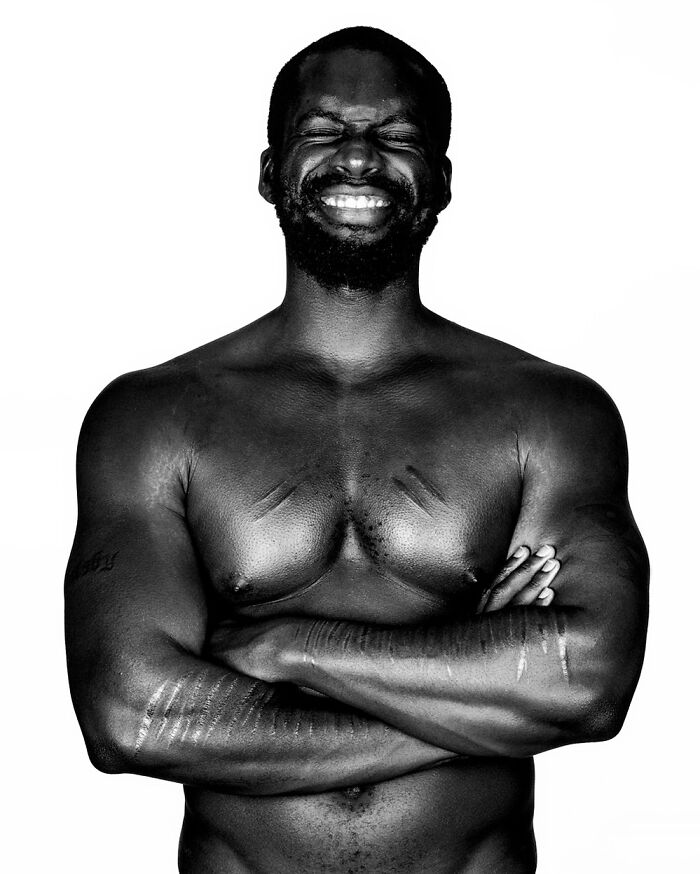 A black and white portrait by Brock Elbank of a muscular man with a beard, smiling with his eyes closed, arms crossed.